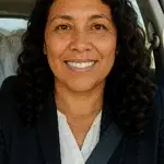 Confident woman smiling while seated in a car, wearing a dark blazer and seatbelt.