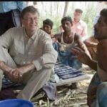 Bill Gates sitting cross-legged on a mat, listening attentively to a local villager in a rural setting.