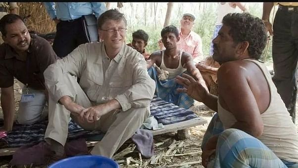 Bill Gates sitting cross-legged on a mat, listening attentively to a local villager in a rural setting.