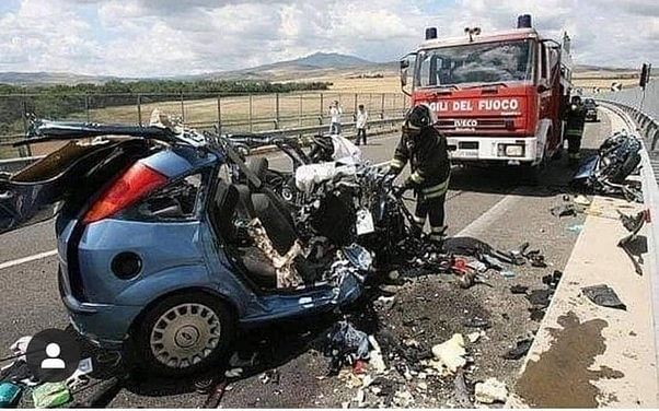 Wrecked blue car on a highway after a deadly crash, with firefighters and a fire truck at the scene