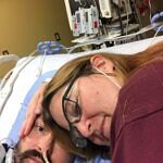 A woman lying beside her husband in a hospital bed, holding his face gently.