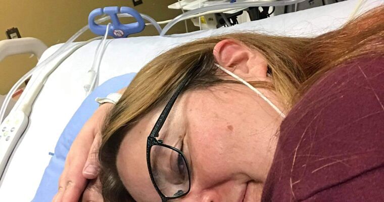 A woman lying beside her husband in a hospital bed, holding his face gently.