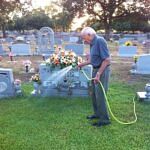 Elderly man watering flowers on a grave in a cemetery at sunset.