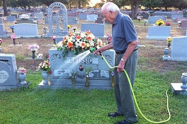 Elderly man watering flowers on a grave in a cemetery at sunset.