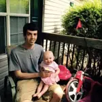 Young man sitting on a porch, holding a smiling baby on his lap.